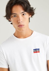White cotton T-shirt featuring a small Levi's logo patch in red, blue, and white on the left chest. Classic design with a round neckline.
