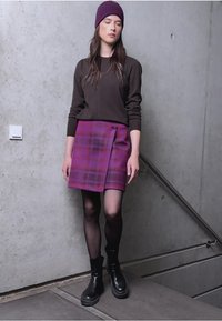 Jupe portefeuille à carreaux violets, chemise à manches longues marron, collants noirs et bottes noires imposantes. Les accessoires comprennent un bonnet violet foncé. Fond en béton.