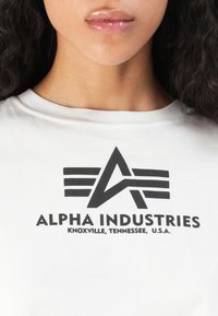 Lähikuva henkilöstä, joka on pukeutunut valkoiseen paitaan, jossa on musta Alpha Industries -logo ja teksti "Knoxville, Tennessee, Yhdysvallat."