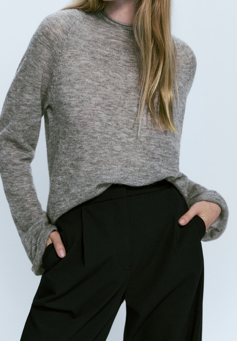 Femme portant un pull gris doux à manches longues rentré dans un pantalon noir taille haute, les mains dans les poches.
