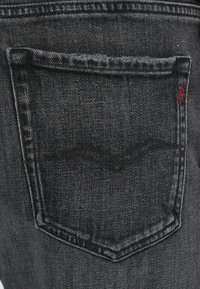 Fekete farmernadrág zsebének finom íves varrási részlete, megerősített varrás és a jobb felső sarokban egy piros logó