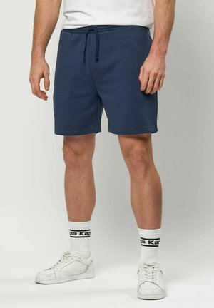 Uomo che indossa pantaloncini blu navy con coulisse, calzini bianchi con logo e scarpe da ginnastica bianche, in piedi davanti a uno sfondo chiaro semplice.