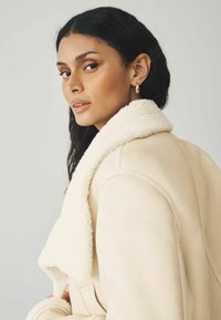 Crème schapenvacht jacket met een zachte kraag en strakke lijnen. Het materiaal lijkt zacht en verhoogt de warmte en het comfort. Sieraden omvatten eenvoudige gouden oorbellen.