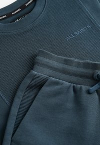 Tamsiai žalias džemperis ir kelnių komplektas. Raukšlėta tekstūra, įspaustas logotipas ant džemperio, elastinga juosmens juosta ir reguliuojama raištelė.