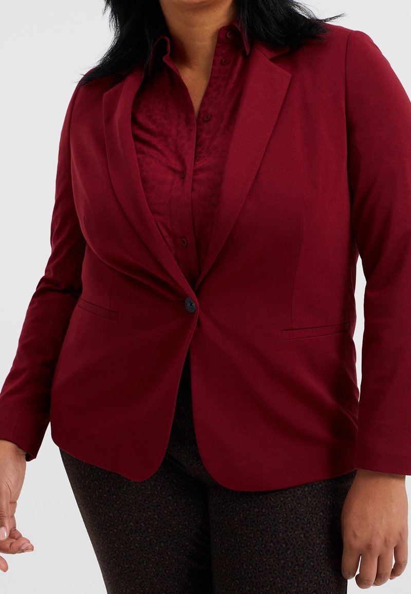 Burgundy blazer with a single black button, smooth fabric, notched lapels, and styled over a patterned burgundy shirt.