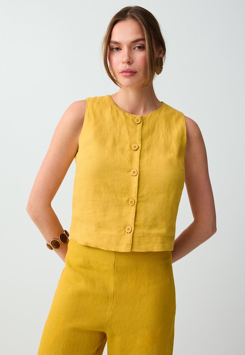 Femme portant un haut jaune moutarde sans manches boutonné et un pantalon taille haute assorti, accessoirisée d'un bracelet et de boucles d'oreilles, sur un fond uni.
