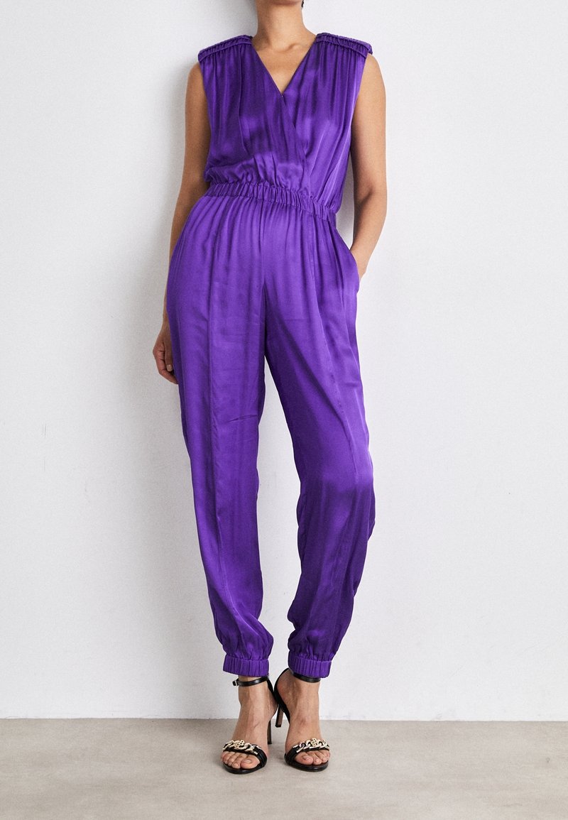 Combinaison en satin violet avec un col en V, une taille élastique et des détails froncés aux épaules. Caractérisée par des jambes fuselées et des poignets.