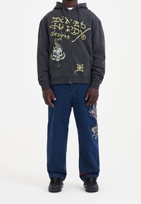 Felpa grigia con zip davanti, caratterizzata da una grafica con teschio e dettagli a forma di serpente, con grandi scritte. Abbinata a jeans blu decorati con un ricamo colorato di un drago.