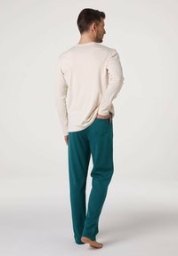 Camicia a maniche lunghe color beige chiaro abbinata a pantaloni lounge verde petrolio. La camicia ha una scollatura rotonda, mentre i pantaloni presentano una tasca posteriore.