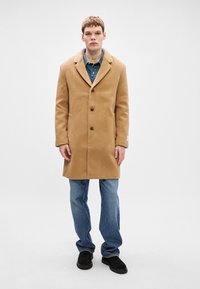 Καμηλί μπεζ μάλλινο παλτό με κομμένο γιακά, τρία κουμπιά και πλαϊνές τσέπες, φορεμένο πάνω από ένα τζιν πουκάμισο και μπλε τζιν.