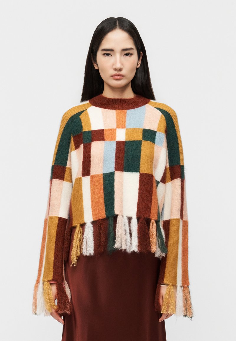 Femme aux longs cheveux noirs portant un pull à carreaux multicolores avec des détails en franges et une jupe marron, debout devant un fond uni.