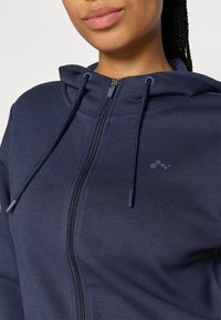 Námořnická mikina na zip vyrobená z měkké tkaniny, s předním zipem, šňůrkami a malým logem na hrudi.