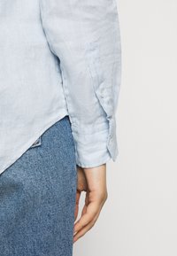 Höger arm och hand på en person som bär en ljusblå långärmad skjorta och blå denimjeans mot en enfärgad vit bakgrund.