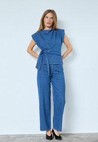 Ținută din denim care include un top drapat fără mâneci, cu o talie înnodată, combinat cu blugi cu picioare largi. Ambele sunt de culoare albastru uniform.