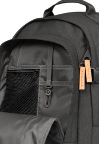 Sac à dos en tissu gris avec une poche en mesh, une fermeture éclair et un crochet à pression. Présente des accents ressemblant à du cuir. Texture lisse et forme arrondie.