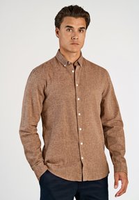 Bruine blouse met lange mouwen en knopen, gemaakt van een gestructureerde stof met een puntige kraag en witte knopen, getoond op een model tegen een effen achtergrond.