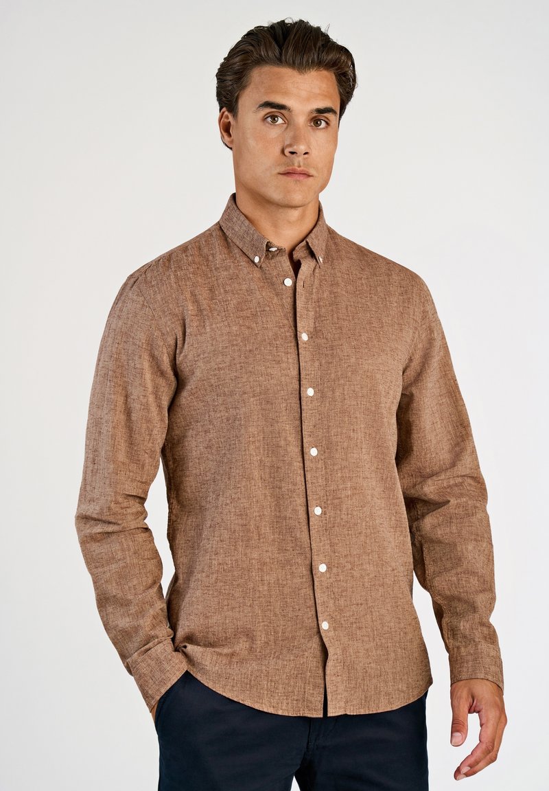 Bruine blouse met lange mouwen en knopen, gemaakt van een gestructureerde stof met een puntige kraag en witte knopen, getoond op een model tegen een effen achtergrond.