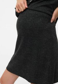Donkergrijze geruite rok gedragen door een persoon met de handen op de taille en een opgevouwen zwarte top zichtbaar aan de bovenrand.