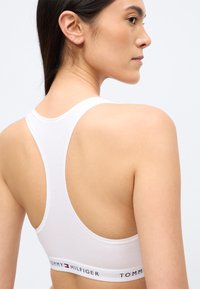 Fehér sportmelltartó racerback kialakítással. Sima textúrájú, és egy márkázott elasztikus szalaggal rendelkezik, amelyen a "TOMMY HILFIGER" logó színes hangsúlyokkal látható.