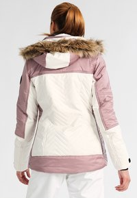 Doudoune blanche et rose pastel, avec capuche en fausse fourrure, texture matelassée et motifs en chevrons dans le dos et sur les manches.