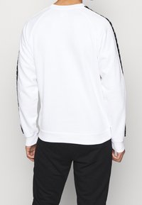 Witte sweatshirt met zwarte zijstrepen, ribbelboorden en onderrand. Zachte textuur, raglanmouwen en een relaxte fit. Zwarte broeken zichtbaar hieronder.