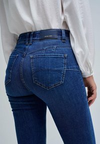 Los jeans de mezclilla azul oscuro presentan un diseño ajustado con bolsillos traseros y una etiqueta de cuero en la cinturilla. La tela muestra un acabado texturizado.