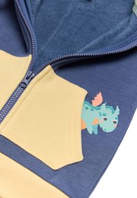 Sudadera con cremallera de diseño bicolor: azul marino y amarillo. Cuenta con un interior suave y un gráfico de un dragón de dibujos animados en el bolsillo.
