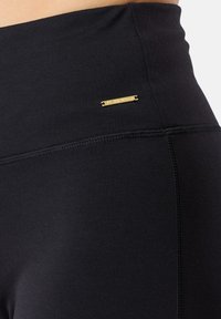 Leggings neri a vita alta realizzati in tessuto elasticizzato, caratterizzati da una leggera lucentezza, cuciture piatte e un piccolo logo dorato.