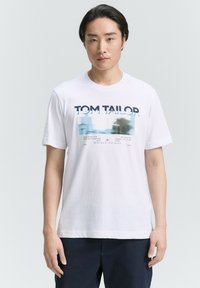 Valkoinen puuvillapaita, jossa on "TOM TAILOR" -teksti ja abstrakti muotoilu sinisellä ja mustalla. Lyhyet hihat ja pyöreä kaula-aukko.