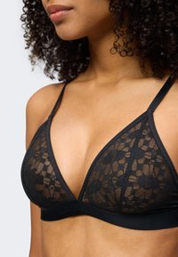 Femme portant un bralette en dentelle noire à fines bretelles et motifs floraux, visible des épaules jusqu'au ventre sur un fond clair.