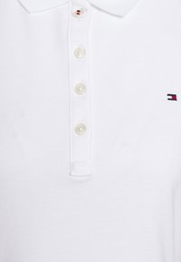 Vit bomullspolo med spetsig krage, sex knappar och en liten flerfärgad logotyp broderad på vänster br öst.