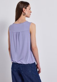 Top senza maniche lilla con dettaglio arricciato sul retro, realizzato in tessuto morbido. Abbinato a pantaloni in denim scuro, che mostrano una vestibilità rilassata.