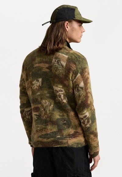 Gebreide trui in groen, bruin en crème met een camouflagepatroon. Inclusief een hoge kraag en geribbelde mouwen. Gepaard met een groene pet.