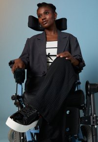 Juodas neįgaliojo vežimėlis su mėlynomis detalėmis; asmuo dėvi pilką švarką, baltą marškinėlį su juodomis juostelėmis ir tekstūruotas juodas kelnes. Juodi blizgūs sportbačiai.