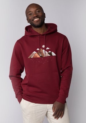 Homme souriant portant un sweat à capuche bordeaux avec un motif de montagne et de soleil, debout avec une main dans la poche sur un fond clair uni.