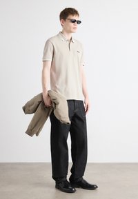 Beige Poloshirt mit hellgrauem Kragen, schwarze Jeans und schwarzen Schuhen. Das Model hält eine beigefarbene Bomberjacke und trägt eine Sonnenbrille. Einfaches, modernes Design.