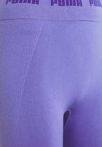Fialové sportovní legíny mají hladkou strukturu s žebrovanými akcenty. Na pásu se výrazně zobrazuje označení "PUMA" v kontrastním designu.
