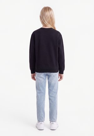 Kind met lang blond haar, van achteren gezien, draagt een zwarte trui, lichtblauwe spijkerbroek en witte sneakers.