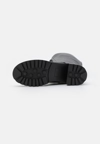 Semelle en caoutchouc noir avec motif de bande de roulement texturé, mettant en avant des accents hexagonaux et rainurés. La partie supérieure est en tissu gris. Le design global de la botte est bas.