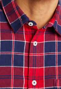 Kołnierz i górna część koszuli w czerwono-granatową kratę z guzikami na osobie z widoczną skórą nad linią szyi.
