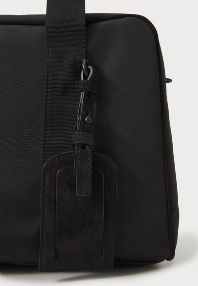 Geantă din material textil negru cu un exterior neted, formă rectangulară și un etichetă din faux suede cusută, prevăzută cu un bucle și închidere cu buton.
