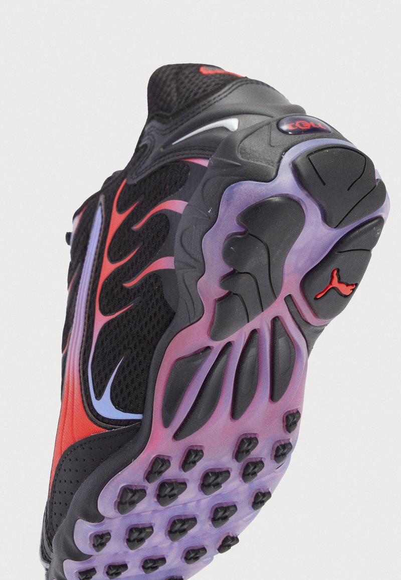 Zapatilla deportiva negra con acentos rojos y púrpuras, parte superior de malla, suela texturizada, surcos pronunciados y un patrón de suela distintivo.