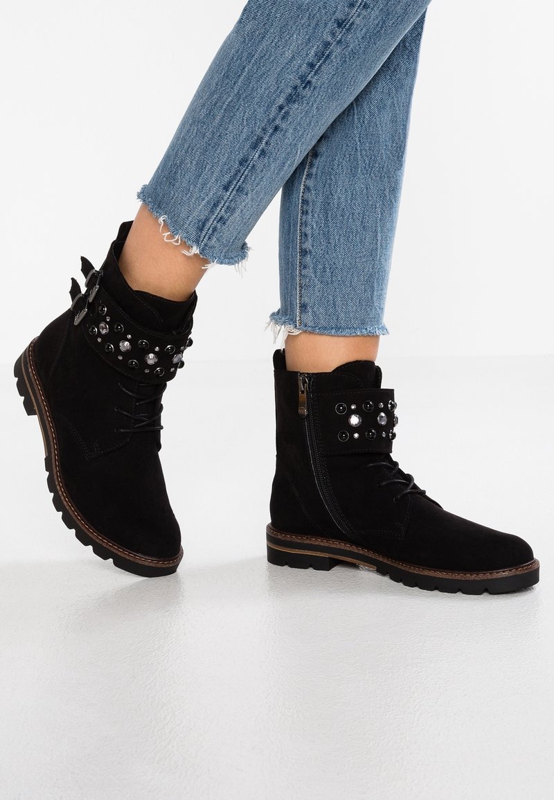 Bottes chevilles en daim noir à lacets sur le devant, avec une bride décorative cloutée, une fermeture éclair latérale, et une semelle robuste. Portées avec un jean bleu à ourlet effiloché.