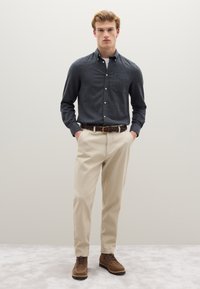 Camisa de botones gris oscuro con mangas largas y un bolsillo frontal, combinada con pantalones chinos beige y zapatos marrones con cordones. Cinturón texturizado con hebilla.