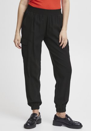 Zwarte joggingbroek van zacht materiaal, met een elastische tailleband, zijzakken en taps toelopende boorden. Gecombineerd met zwarte schoenen.