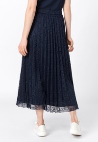 Tmavě modrá plisovaná midi sukně z krajek s texturovaným květinovým vzorem, elastickým pasem a vrstvami krajkového lemu. Pro kontrast jsou nošeny bílé tenisky.