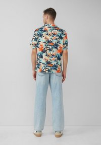 Kurzärmliges, mehrfarbiges Hawaii-Hemd mit Blumen- und Palmenmustern, kombiniert mit hellblauen, weit geschnittenen Jeans und beigefarbenen Schuhen.