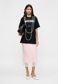 Nő fekete Moschino pólóban, hosszú gyöngysorokkal, rózsaszín csipkével díszített szoknyában, fehér magassarkú cipővel, fekete táskát tartva.