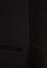 Zwarte stof met een zachte textuur en een subtiele glans. Voorzien van een paspelzak met duidelijke stiksels en zichtbare naden.