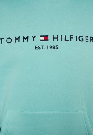 Tissu bleu clair avec texte brodé "Tommy Hilfiger" et logo en rouge, blanc et bleu, avec "EST. 1985" sous le logo.